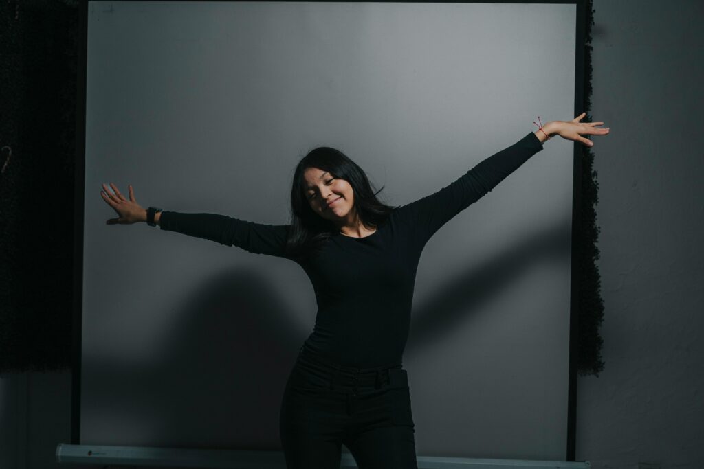 A woman joyfully stretches her arms wide in a dimly lit room.